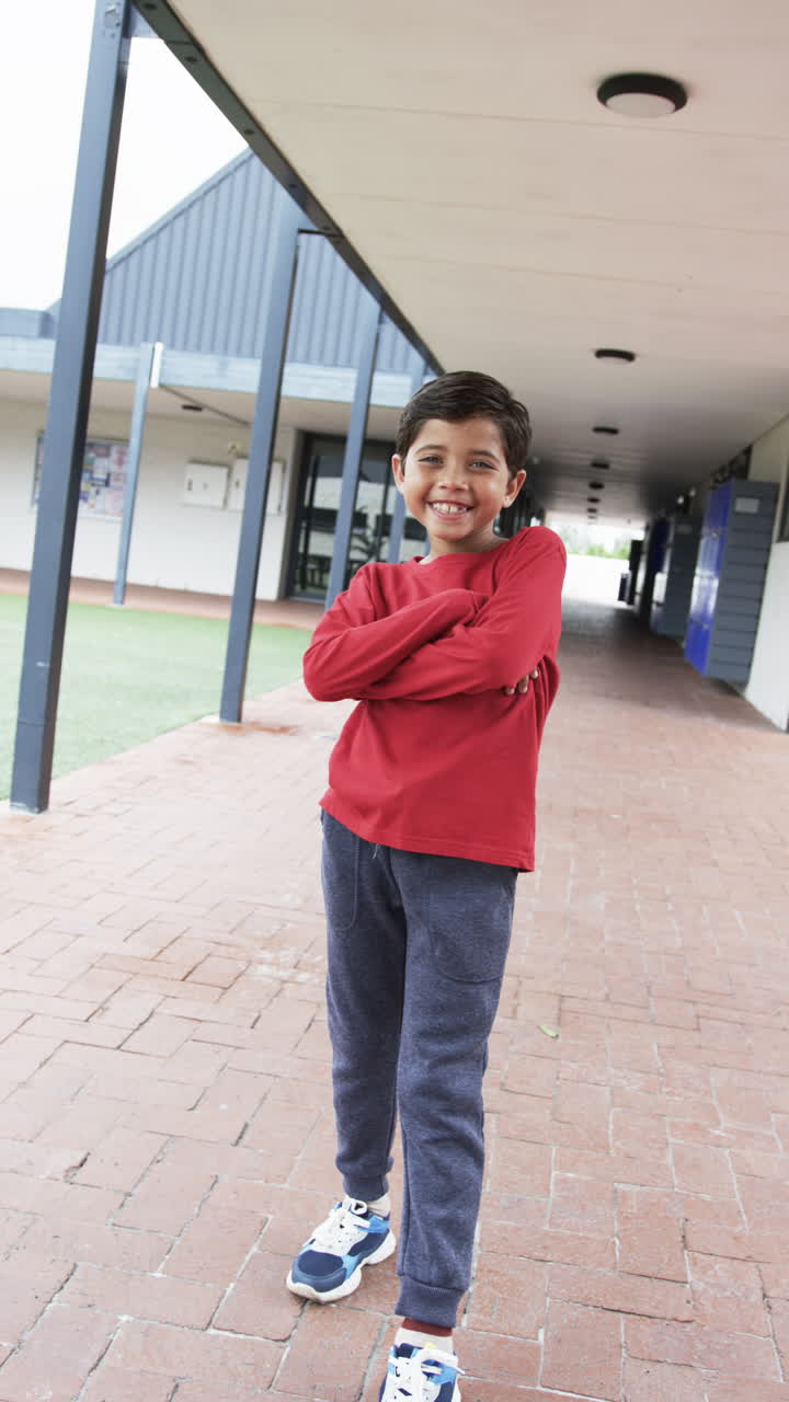Vertical video: In a school corridor, a young Caucasian boy stands with arms crossed