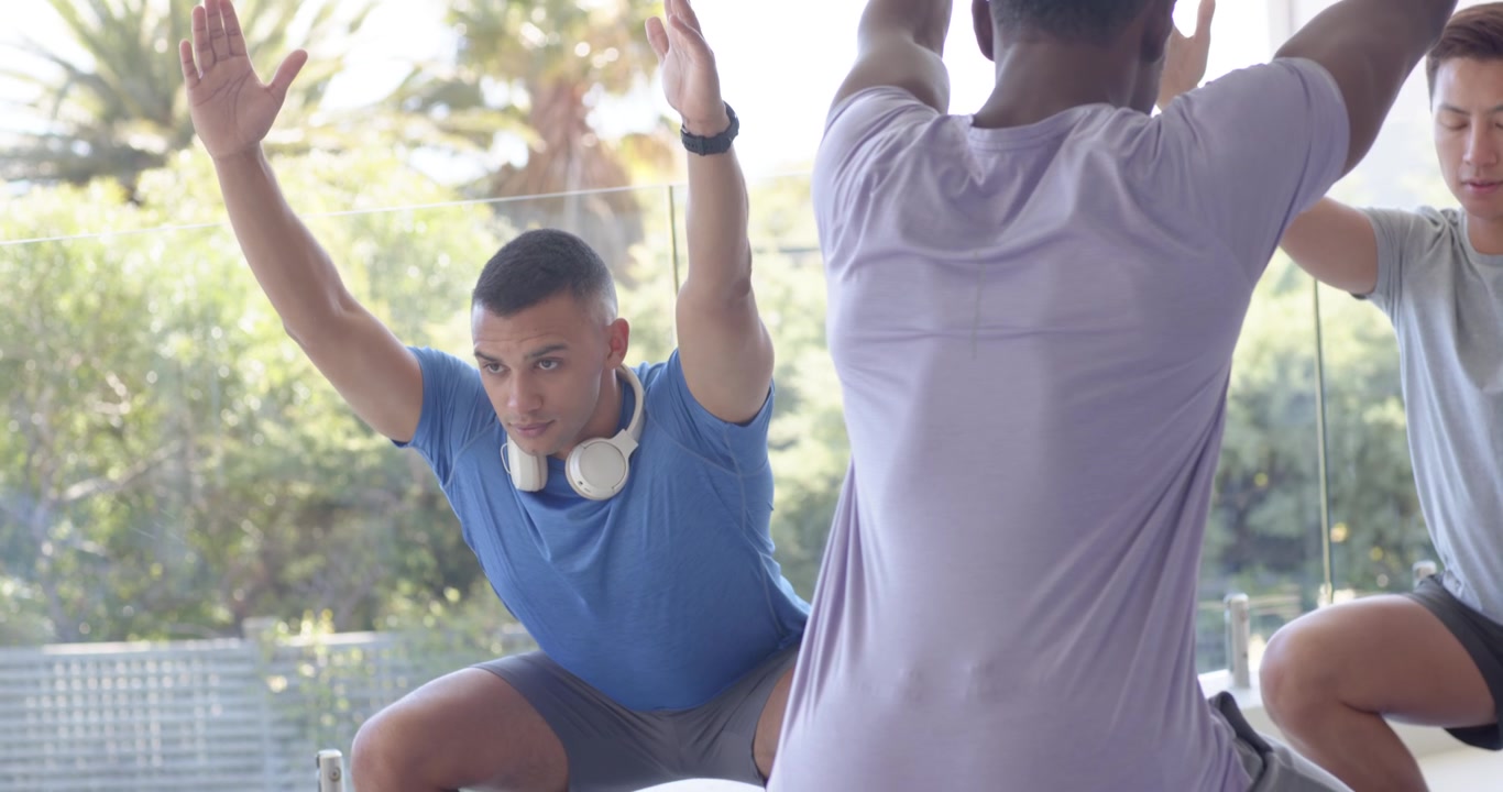 Uomini sportivi di diverse etnie che seguono un leader centrale alzando le braccia, imitando gli squat in una palestra di vetro con cuffie