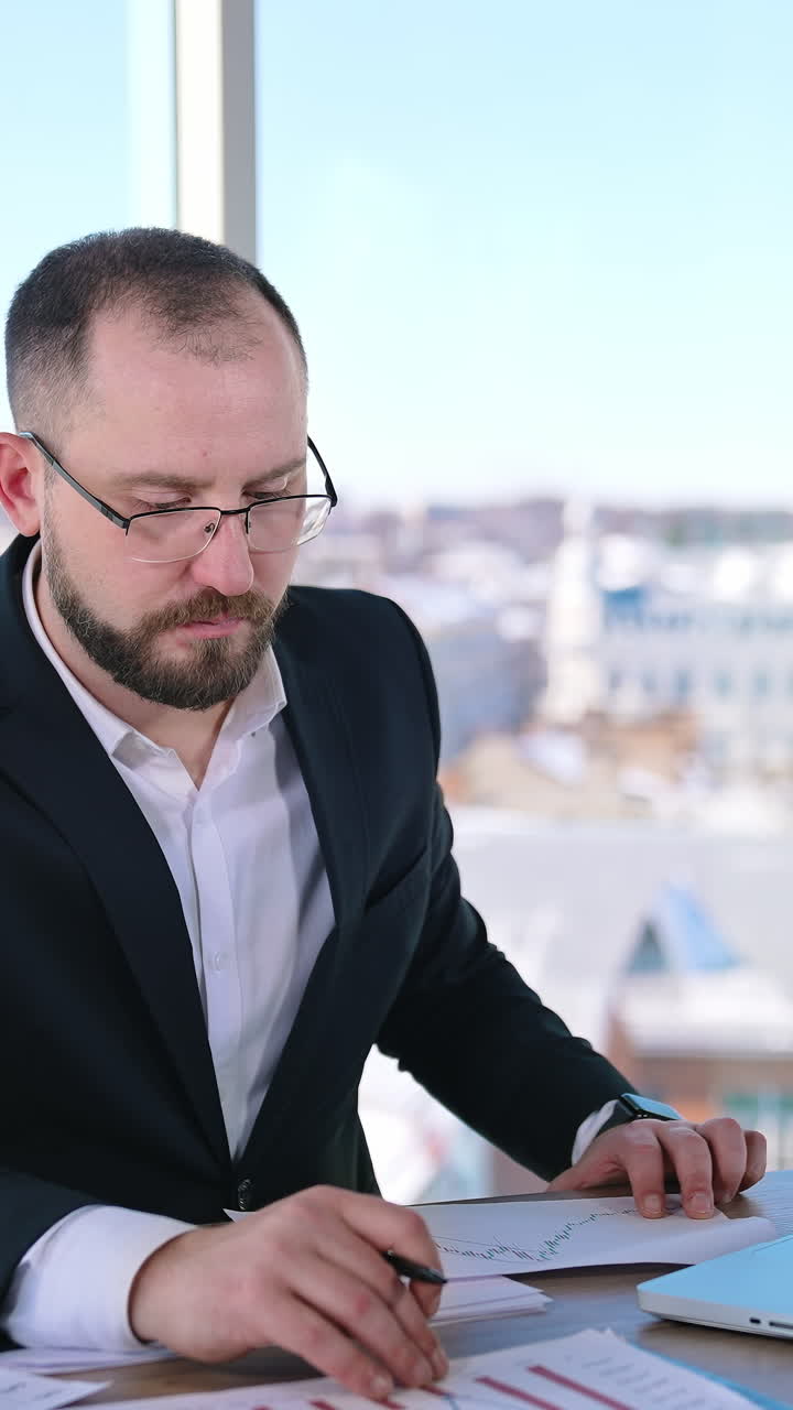 Portrait of a serious office worker indoors. Businessman in glasses sitting at desk and working on new project in office near the window. Vertical video