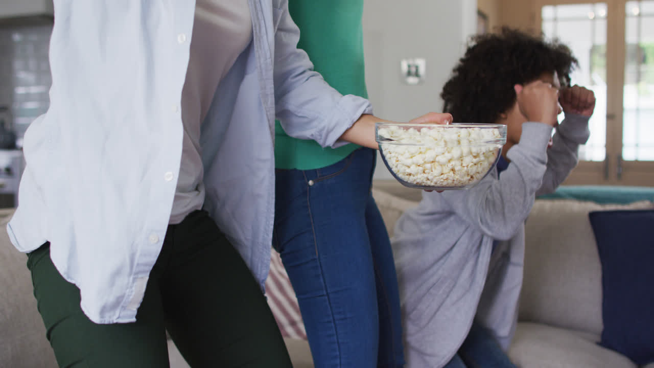 una pareja de lesbianas de raza mixta y su hija viendo la televisión comiendo palomitas de maíz.