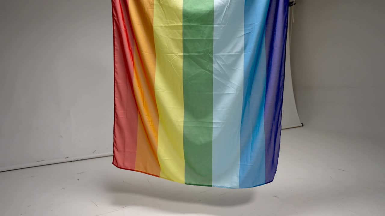 Bandeira de arco-íris se assentando e deslocando-se para a direita e para baixo no estúdio pela gravidade, revelando dobras e uma presilha.