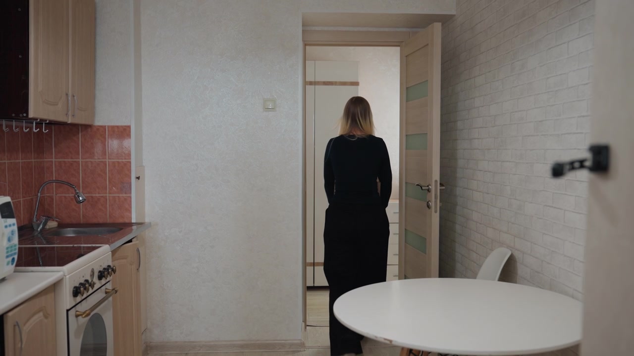 Donna che porta una scatola fuori dalla cucina, lasciando l'appartamento in affitto dopo il trasloco, piani di lavoro vuoti, silenzio echeggiante, un ultimo sguardo, chiude a chiave la porta, chiudendo un capitolo, transito attraverso il corridoio, luce del tardo pomeriggio, riflessiva.