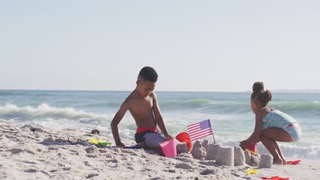 smiling african american siblings building sandcastle with american flag on sunny beach