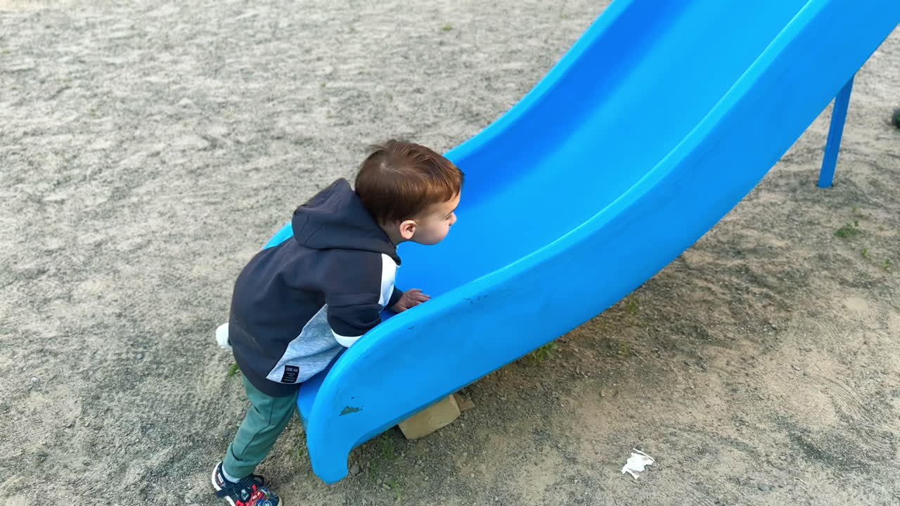Caucasian toddler in hoodie walks up to the slide. Baby boy tries to climb up by the slide but goes down.