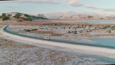 Aerial-view-following-vehicle-on-Reynisfjara-main-road-travelling-across-snowy-Icelandic-black-sand-landscape