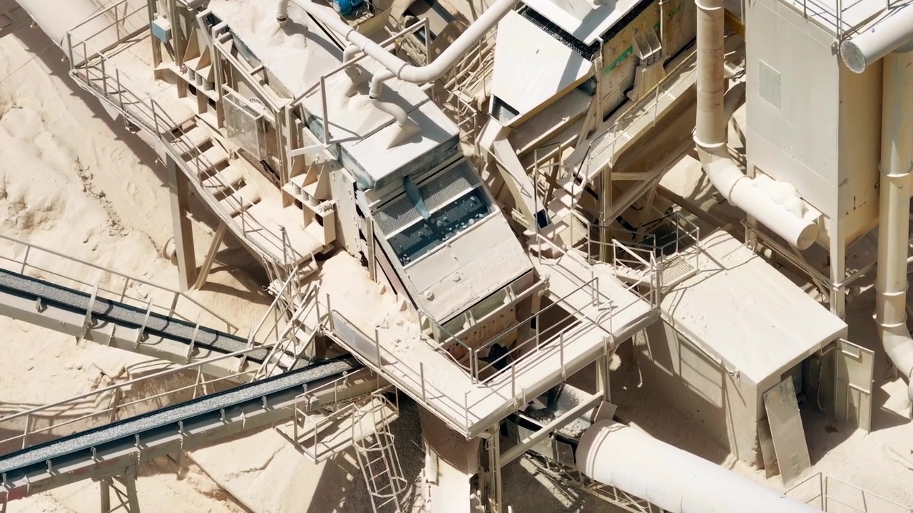 Aerial view of a cement production facility. Heavy machinery moves limestone and aggregates. Conveyor belts transport raw materials for processing. Quarry logistics support construction materials.