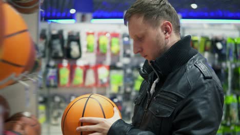 An-adult-man-in-black-leather-jacket-examines-the-balls-that-lie-on-the-shelves-in-the-supermarket.-A-man-looks-at-a-basketball-ball-for-outdoor-sports