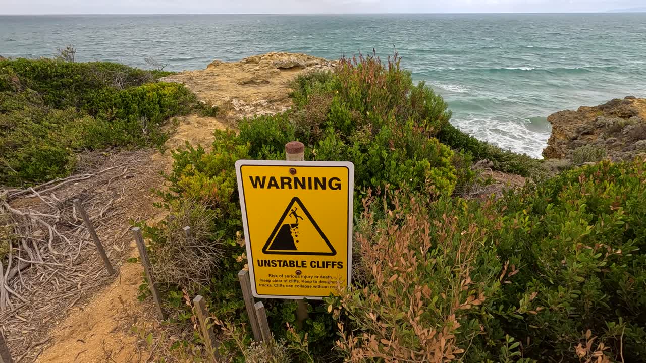 A handheld camera slowly pans across a coastal cliffside with a yellow warning sign, natural vegetation, and ocean backdrop in daylight