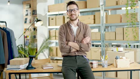 cucasian designer man in eyeglasses leaning on the table and looking at camera in a good mood in a fashion clothing store