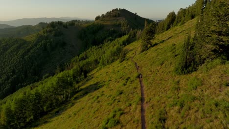 aerial over backcountry mountain range as two bikers bike down trail at sunset-1