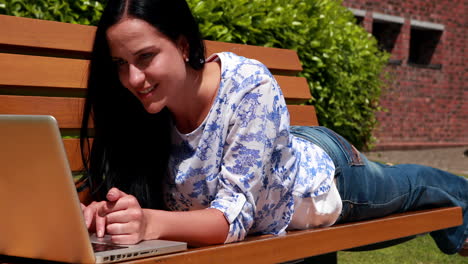 Woman-lying-on-park-bench-using-laptop