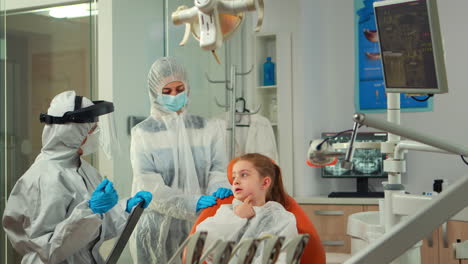 Médico-Dentista-Con-Traje-De-Ppe-Tomando-Notas-En-El-Portapapeles-Sobre-Un-Paciente-Pequeño