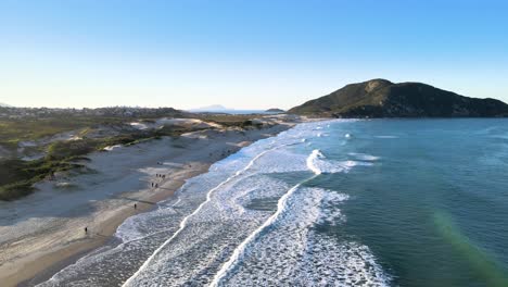 Aerial-drone-view-flying-over-the-beach-with-people-walking-in-the-late-afternoon-with-waves,-dunes-and-hills-montains-revealed-to-the-horizon