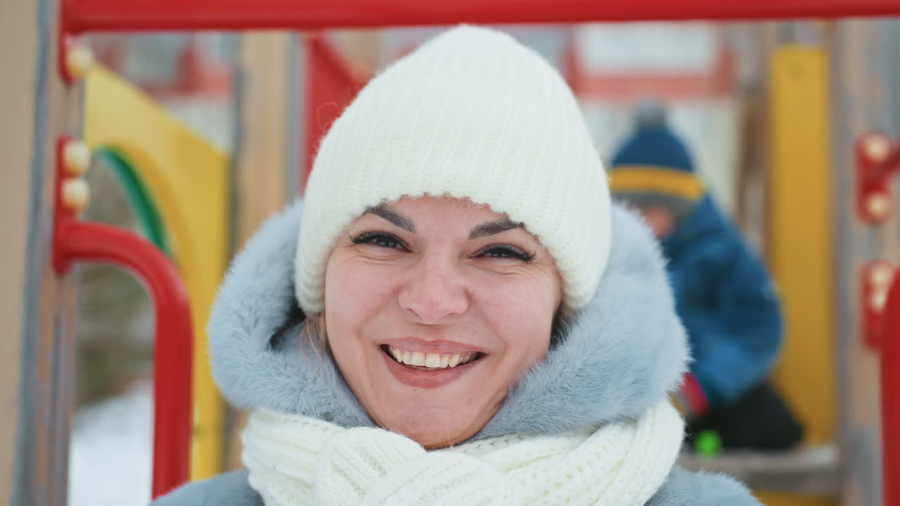 Mamma e figlio sorridenti al parco giochi in primo piano, donna con cappello e sciarpa di lana bianchi, bambino sfocato che gioca su attrezzature colorate, espressioni gioiose e sincere, scena all'aperto innevata, calore e legame familiare.