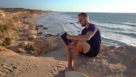 a young man in black dress and sunglass sitting near a rocky beach with ocean in the background at hadera, israel