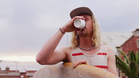 caucasian male surfer holding a wooden surfboard and drinking coffee