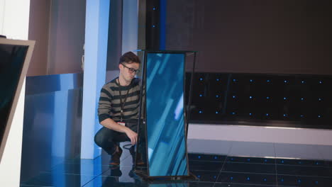 man adjusts display screen setup in video production studio, focused on connecting cables, with camera on tripod and blue lighting, preparing studio space for filming and broadcasting