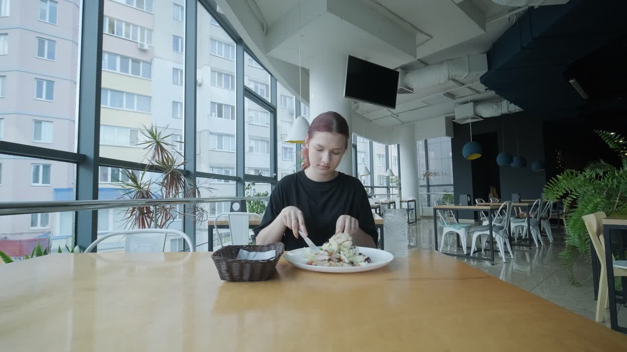 mujer joven comiendo una ensalada fresca en una mesa de madera en un restaurante moderno con grandes ventanas, luz natural y vistas urbanas. escena de comedor relajada