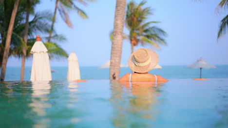 close-up of the back and shoulders of a woman wearing a straw sun hat while relaxing on the edge of a resort infinity-edge pool looking out at the beach and ocean beyond