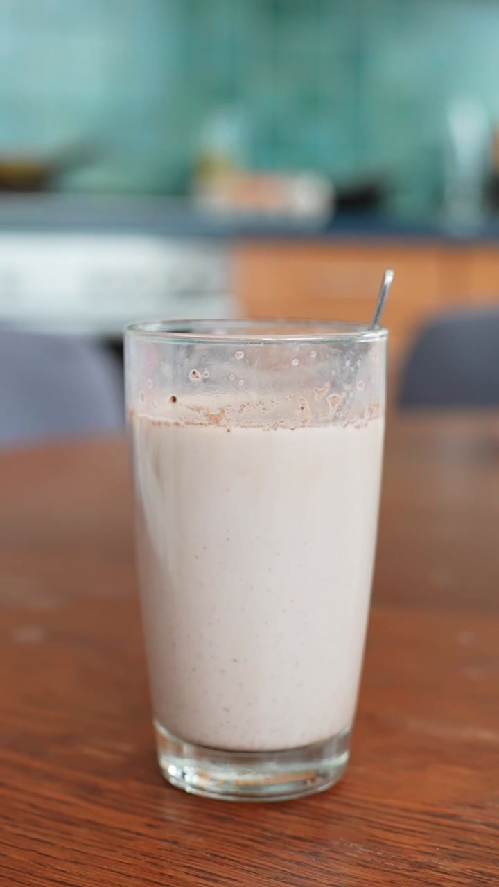 Tall glass of homemade chocolate milk with a spoon on a wooden table. Creamy cocoa drink ready to serve, soft background and copy space. Vertical close up.