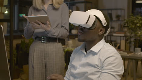 close up view of american man sitting at desk in the office wearing virtual reality glasses while a woman employee is controlling him with tablet