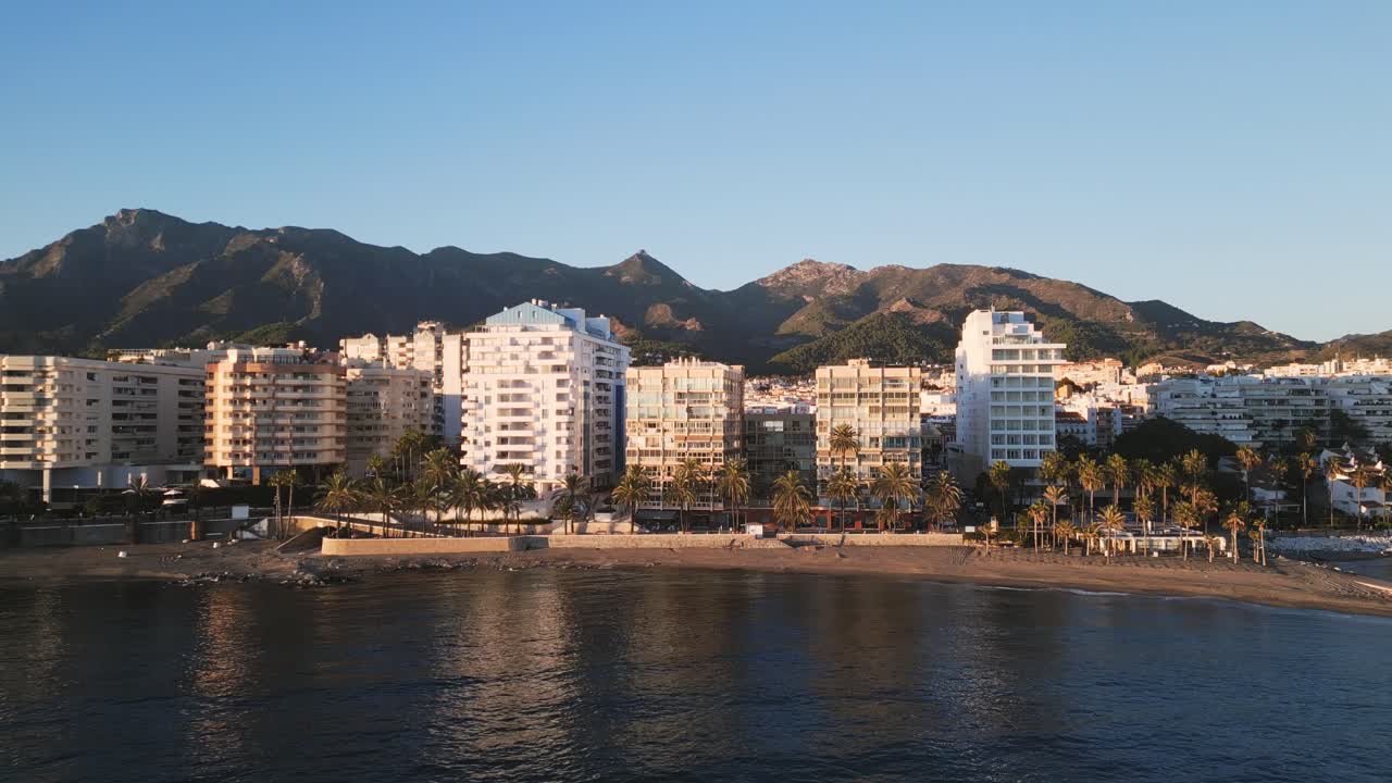 Splendida vista aerea frontale della costa di Marbella con mare calmo in primo piano, skyline della città a metà inquadratura, montagne della Sierra Blanca e cielo blu sopra, riflesso del sole sugli edifici