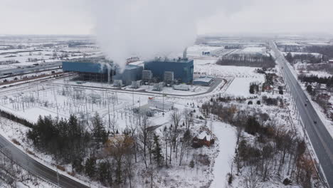 Una-Central-Eléctrica-Industrial-En-Un-Paisaje-Nevado-Con-Chimeneas-Activas,-Carreteras-Y-árboles,-Vista-Aérea