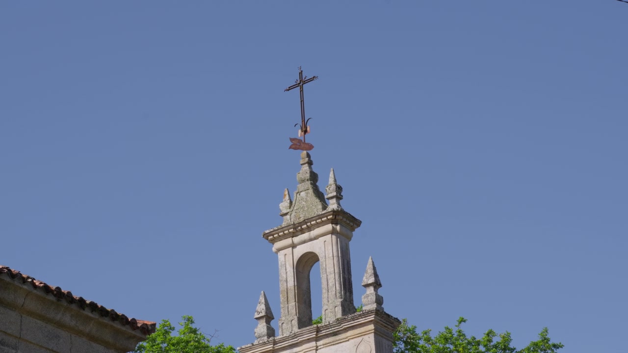 Una ripresa statica media di un campanile di chiesa in pietra ornato sormontato da una croce, sullo sfondo di un cielo blu vibrante e cime di alberi verdi lussureggianti sotto la luce solare intensa.