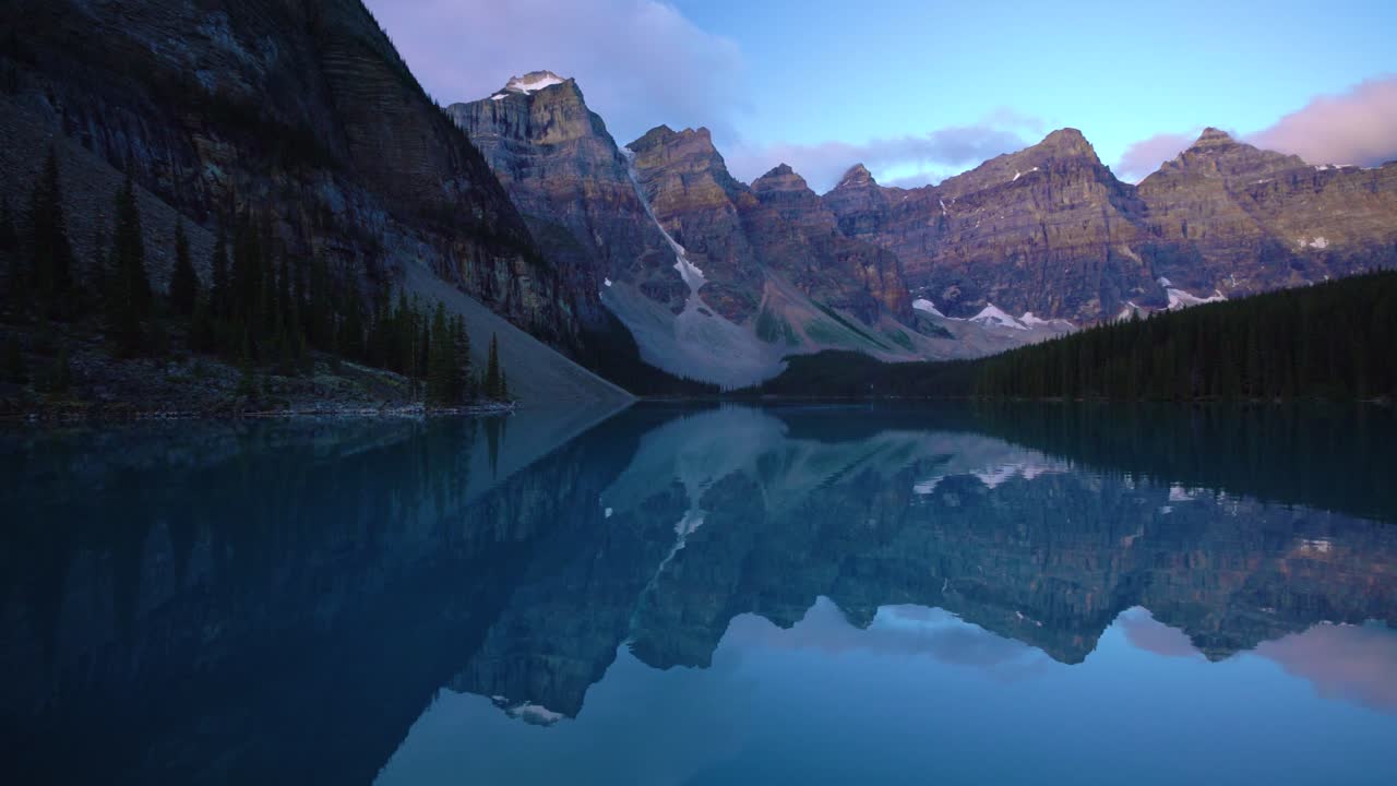 Calm lake reflects towering mountains under soft evening sky in Banff National Park. Pine trees line shore as light fades, creating peaceful natural scene with crystal clear water and rugged peaks