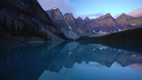 calm lake reflects towering mountains under soft evening sky in banff national park. pine trees line shore as light fades, creating peaceful natural scene with crystal clear water and rugged peaks