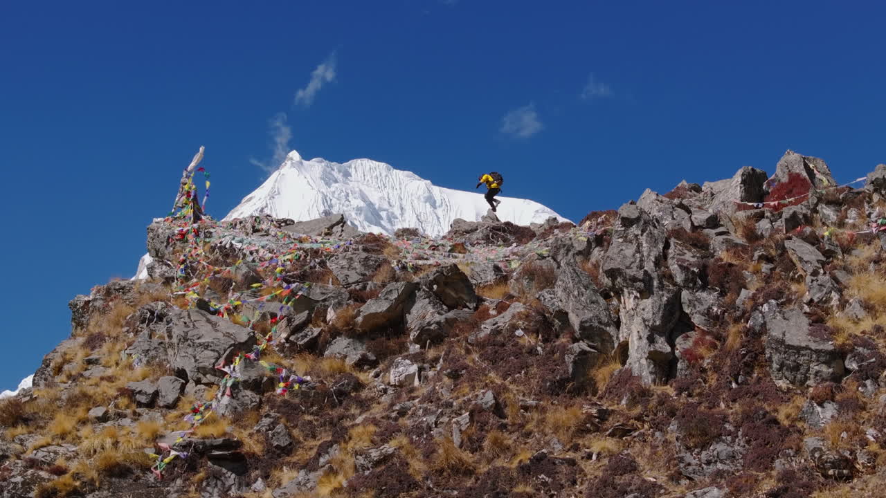 Drone shot reveals a male climber reaching the summit in Langtang, Nepal. Vibrant scene shows snow capped mountains, including Everest, under a clear blue sky, with fluttering prayer flags.