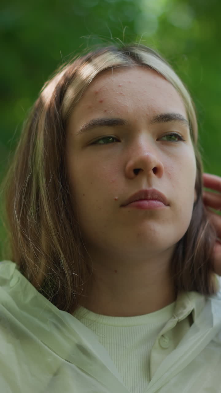 joven con impermeable translúcido mira pensativo, aparentemente ajustando su postura en un entorno al aire libre sereno y verde, su expresión es reflexiva, perdida en pensamientos