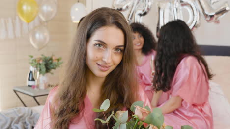 brunette woman holding and smelling a bouquet, wearing pink silk nightdress, smiling and looking at camera