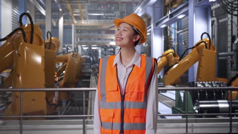 asian female engineer with safety helmet looking around while standing with industrial robotic arms working