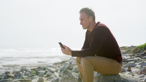 Caucasian-man-enjoying-free-time-by-sea-on-sunny-day-sitting-on-smartphone