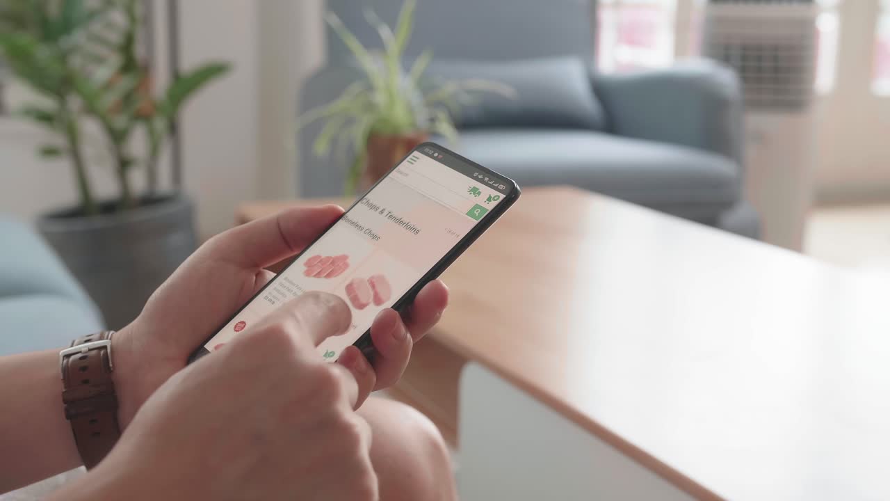 Man Sitting On Couch Using Smartphone Buys In Internet Shop. Male Selects Beef In Grocery Online Store