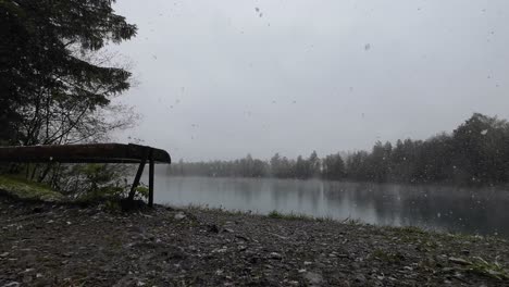 big snow flakes are falling on bench with view over mystical lake in spring