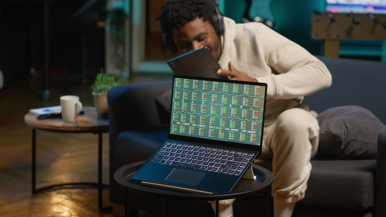 African american person using his tablet laying on the couch while he pays attention to laptop with stock market data trends. Focused man comparing trading graphs numbers before investing. Camera A.