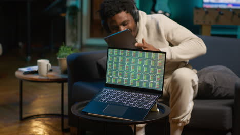 african american person using his tablet laying on the couch while he pays attention to laptop with stock market data trends. focused man comparing trading graphs numbers before investing. camera a.