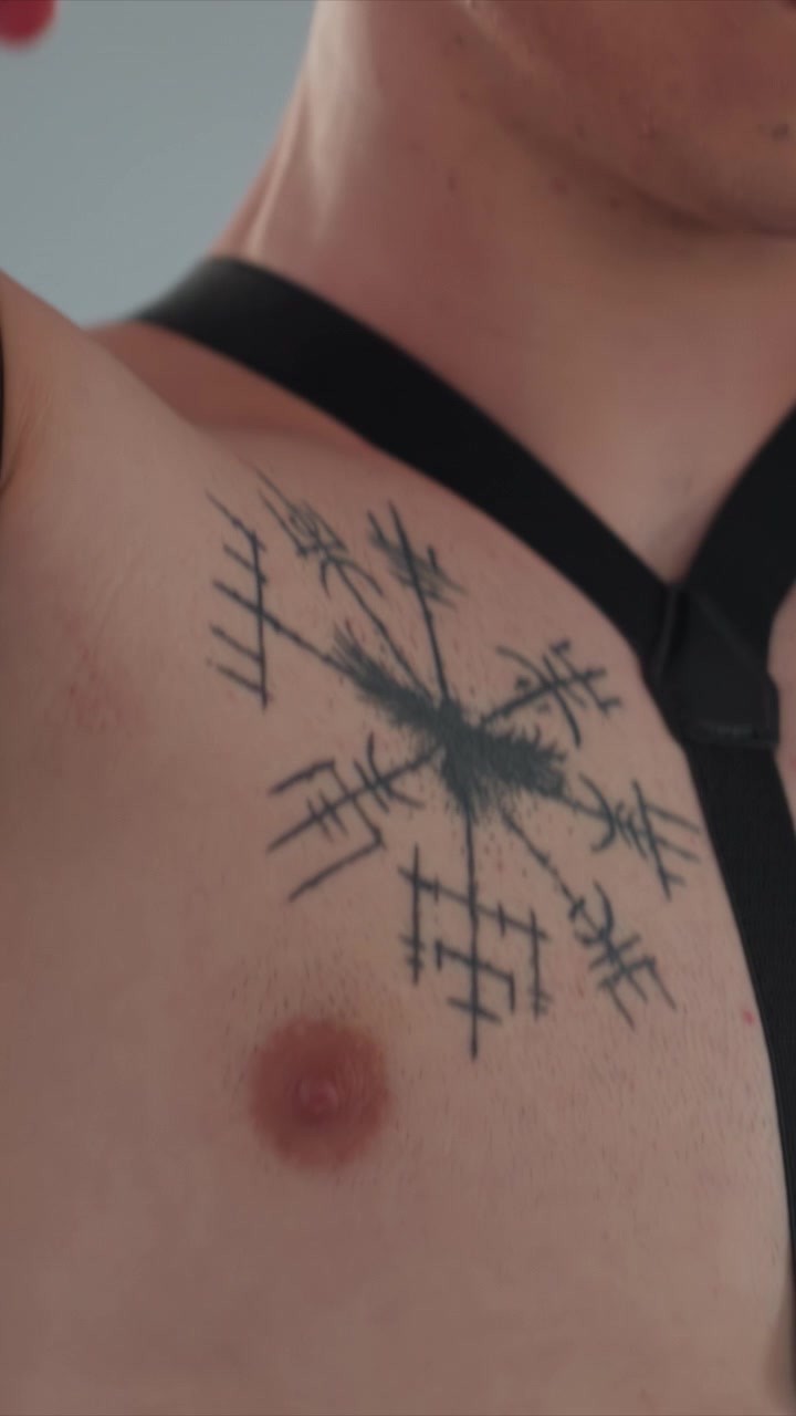 Homem sem camisa com tatuagem no peito ajusta suas calças pretas. O braço de outro homem ajuda a ajustar uma tira na área da cintura. Vista em close-up destaca textura da pele, tatuagens e pintas