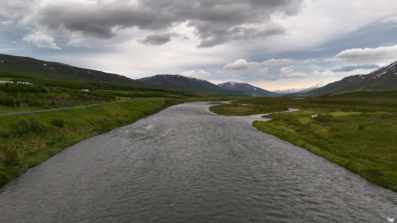 Aerial view of the Skjálfandafljót River winding toward Skjálfandi Bay, framed by broad green valleys, scattered farms and snow-marked mountains under a dramatic sky in northern Iceland