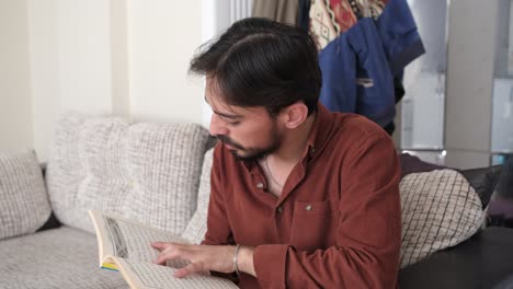 muslim young man sitting on the sofa in his room reading the pages of the quran book, people follow the rules of their religion