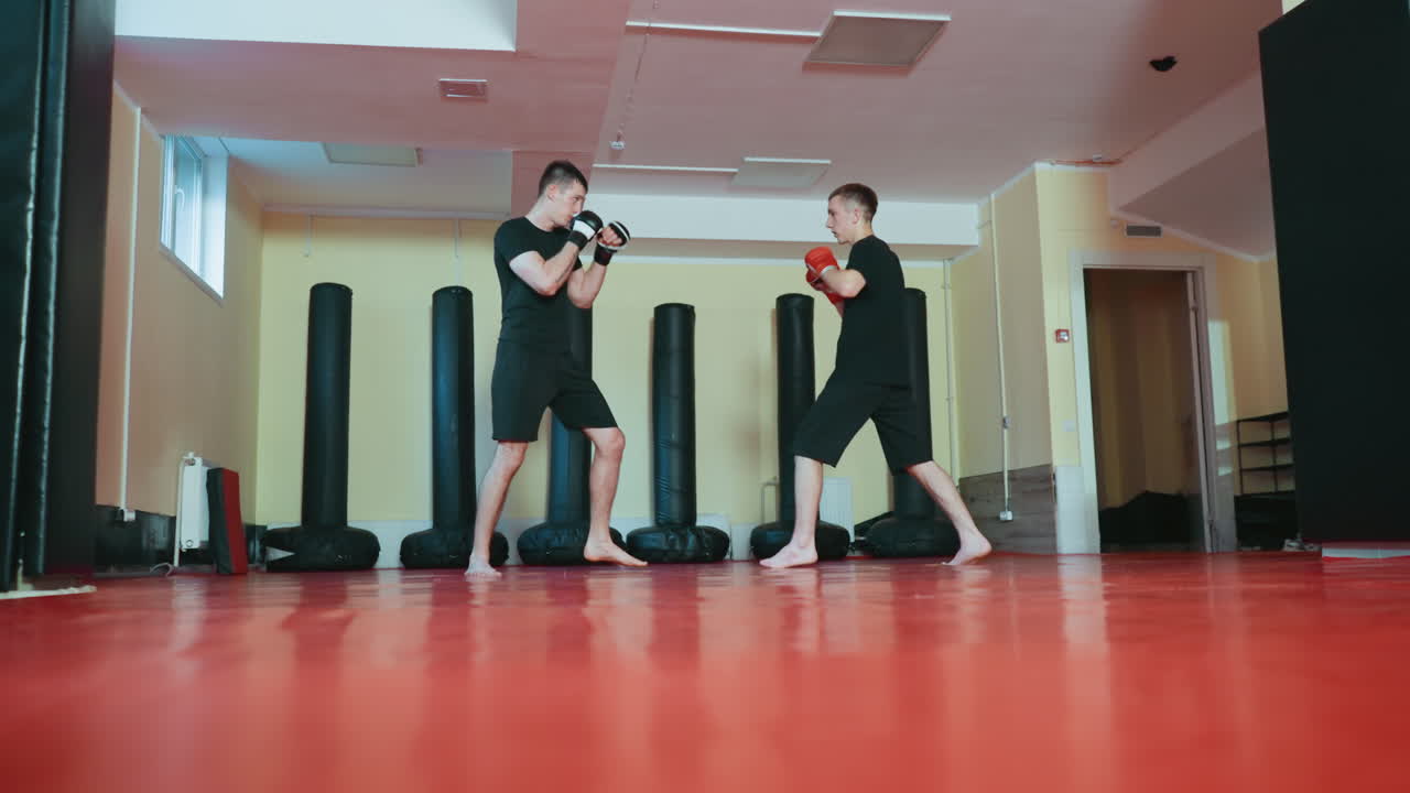 붉은색 매트 위 무술 체육관 안에서 글러브와 검은색 운동복을 입고 서로 마주 보며 격투 자세를 취하고 샌드백으로 둘러싸인 채 전투 훈련 기술을 연습하는 운동선수들의 스파링 모습이에요.