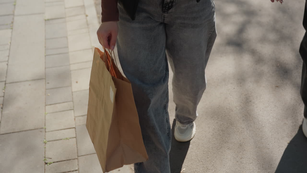persona con ropa vaquera deja una bolsa de papel, individuo con zapatillas deportivas coloca una bolsa de papel sobre listones de madera en un parque, comprador casual vestido con vaqueros y zapatillas deportivas acomoda una bolsa de papel en un banco de madera en un parque