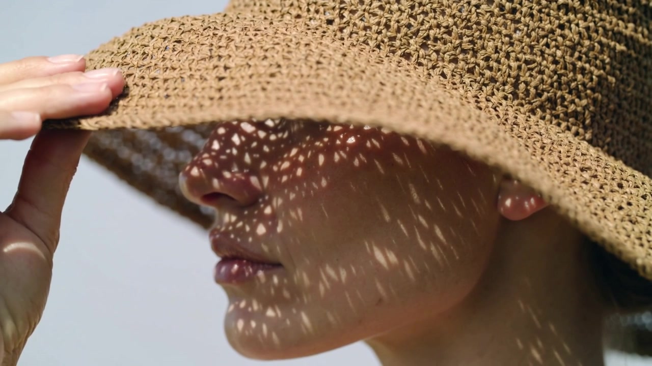 Donna che, afferrando la tesa, abbassa un cappello di paglia sotto un cielo pallido, riparandosi gli occhi sotto una luce screziata