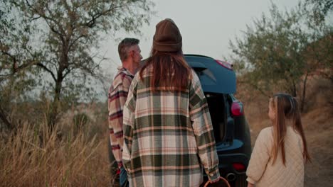 rear view of a brunette girl in a hat in a green checkered shirt, together with her middle-aged husband in a checkered shirt and little daughter, go to a black car and put their things there after a picnic in a summer evening outside the city