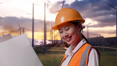 close up side view of asian female engineer with safety helmet looking at blueprint in her hands with solar panel and wind turbines