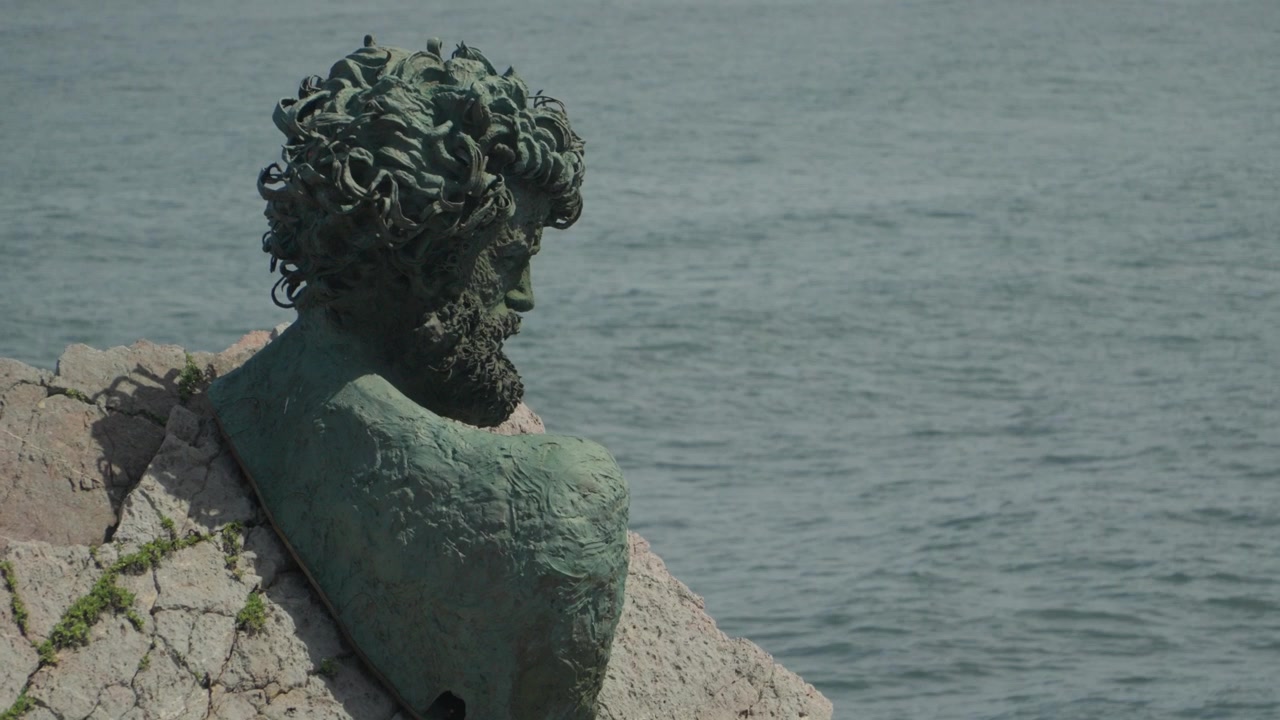 Ampia inquadratura del monumento a busto di Philippe Cousteau integrato nelle scogliere rocciose presso il Museo dell'Ancora a Salinas, Asturie, Spagna. Presenta onde oceaniche che si infrangono e una piattaforma panoramica elevata.