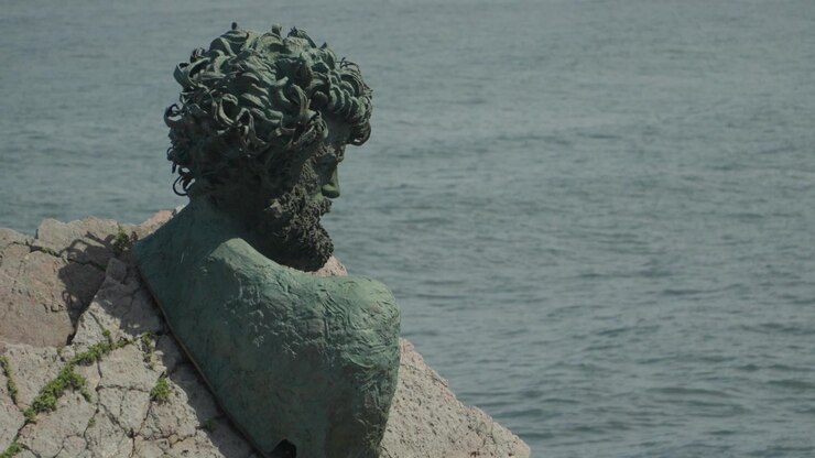 Ampia inquadratura del monumento a busto di Philippe Cousteau integrato nelle scogliere rocciose presso il Museo dell'Ancora a Salinas, Asturie, Spagna. Presenta onde oceaniche che si infrangono e una piattaforma panoramica elevata.