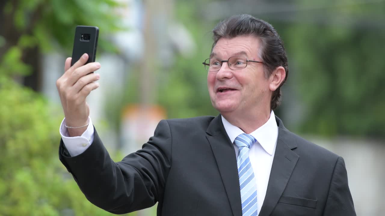 Happy handsome senior businessman video calling in the streets outdoors